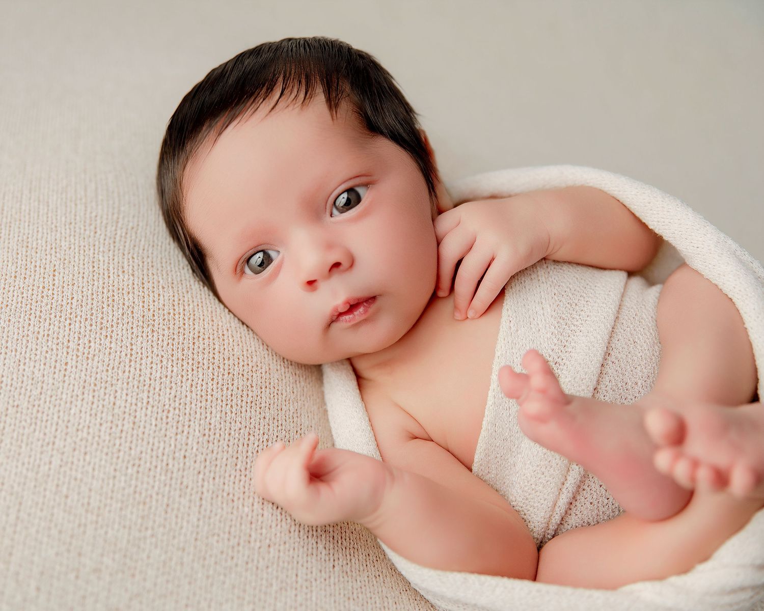A tiny newborn baby wrapped in soft white fabric lies on a beige textured surface while looking at the camera.