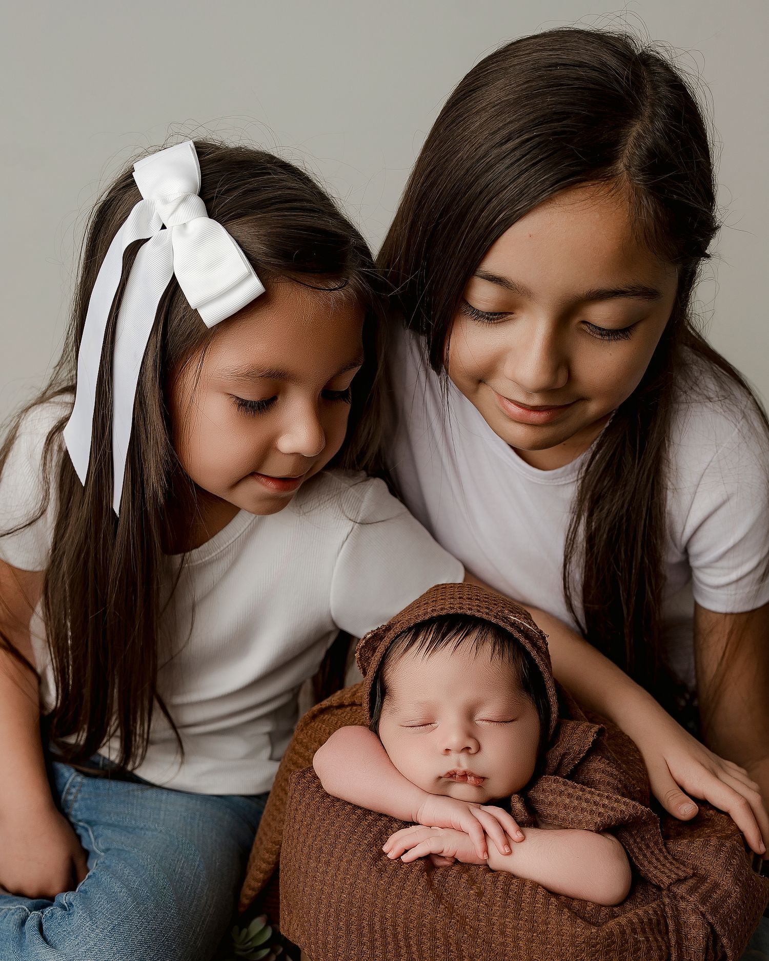 Two siblings in white shirts lovingly watch over a sleeping newborn baby wrapped in a brown blanket.