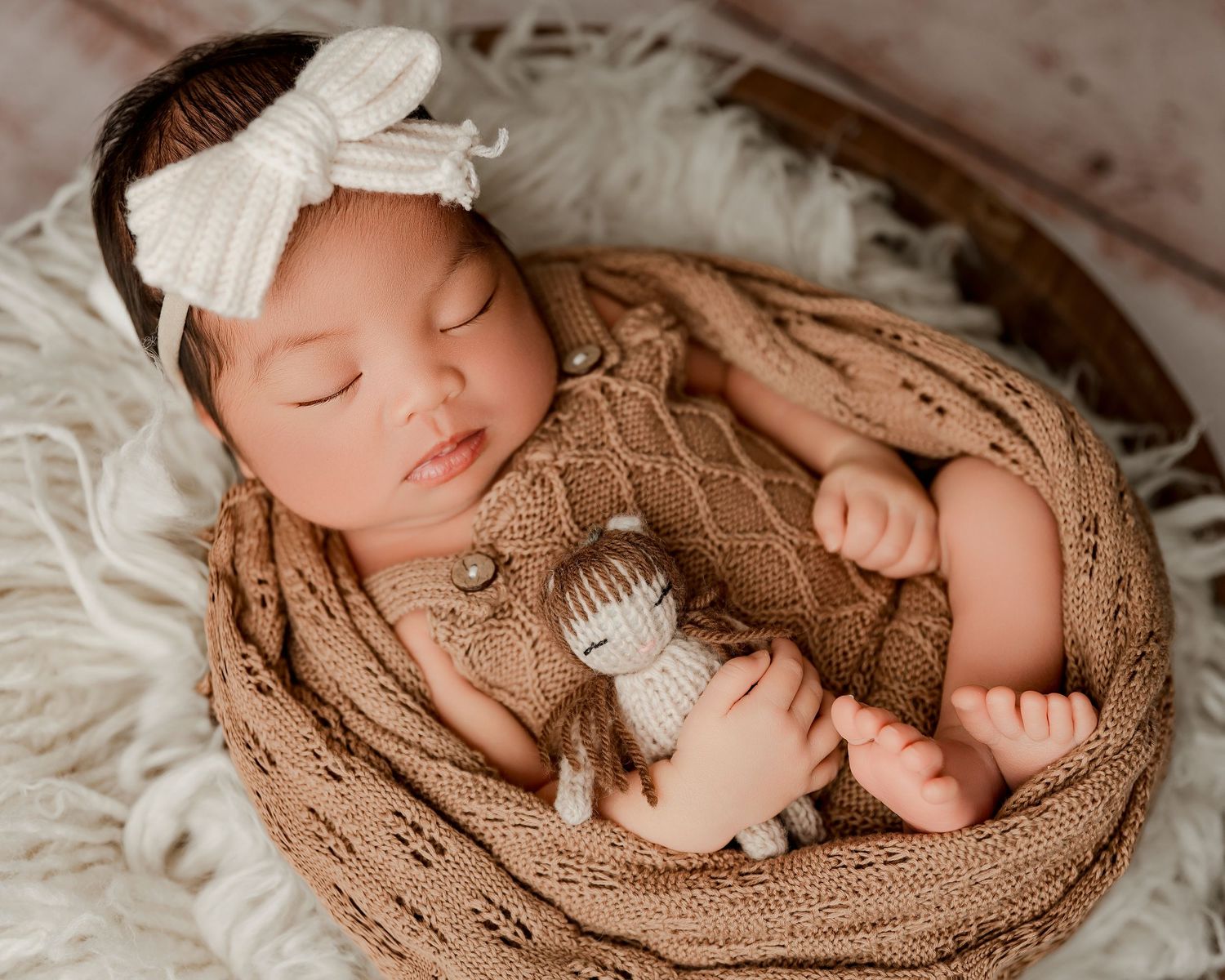 Peaceful newborn sleeping on soft brown knitted blanket with white bow headband in professional portrait setting.