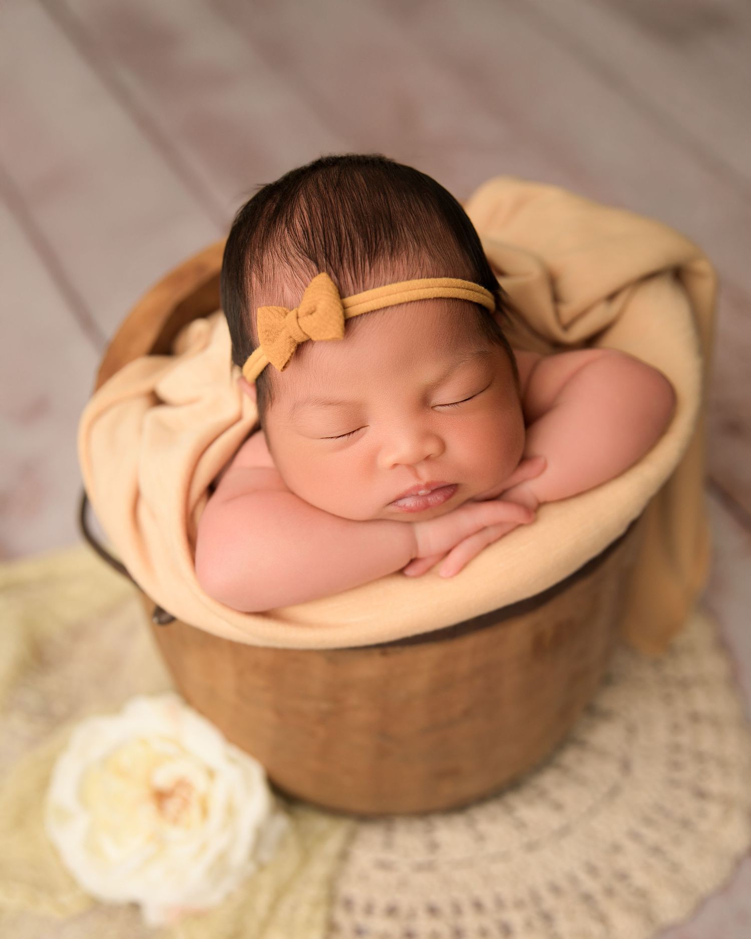 A sleeping newborn baby rests peacefully in a wooden bowl on a textured cream-colored blanket.