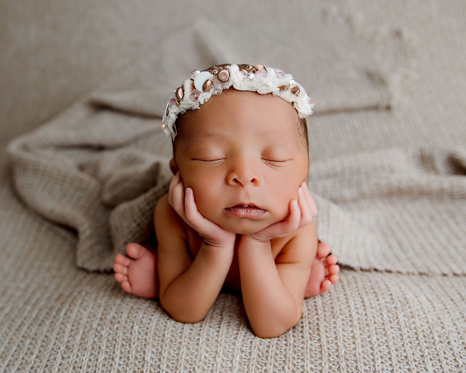 Newborn with floral crown resting head on hands against gray textured backdrop.