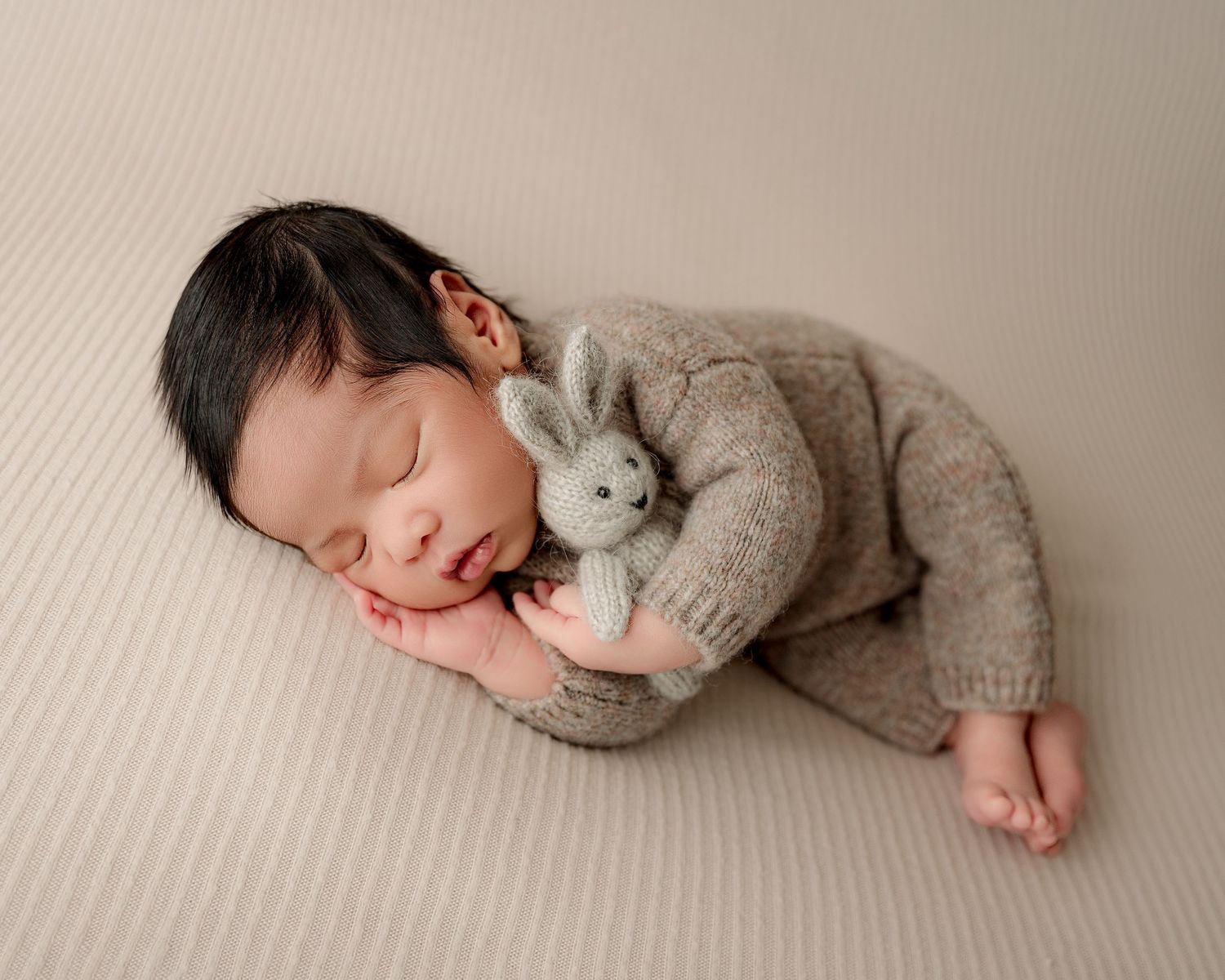 Sleeping newborn in soft grey sweater holding plush bunny toy on textured backdrop.