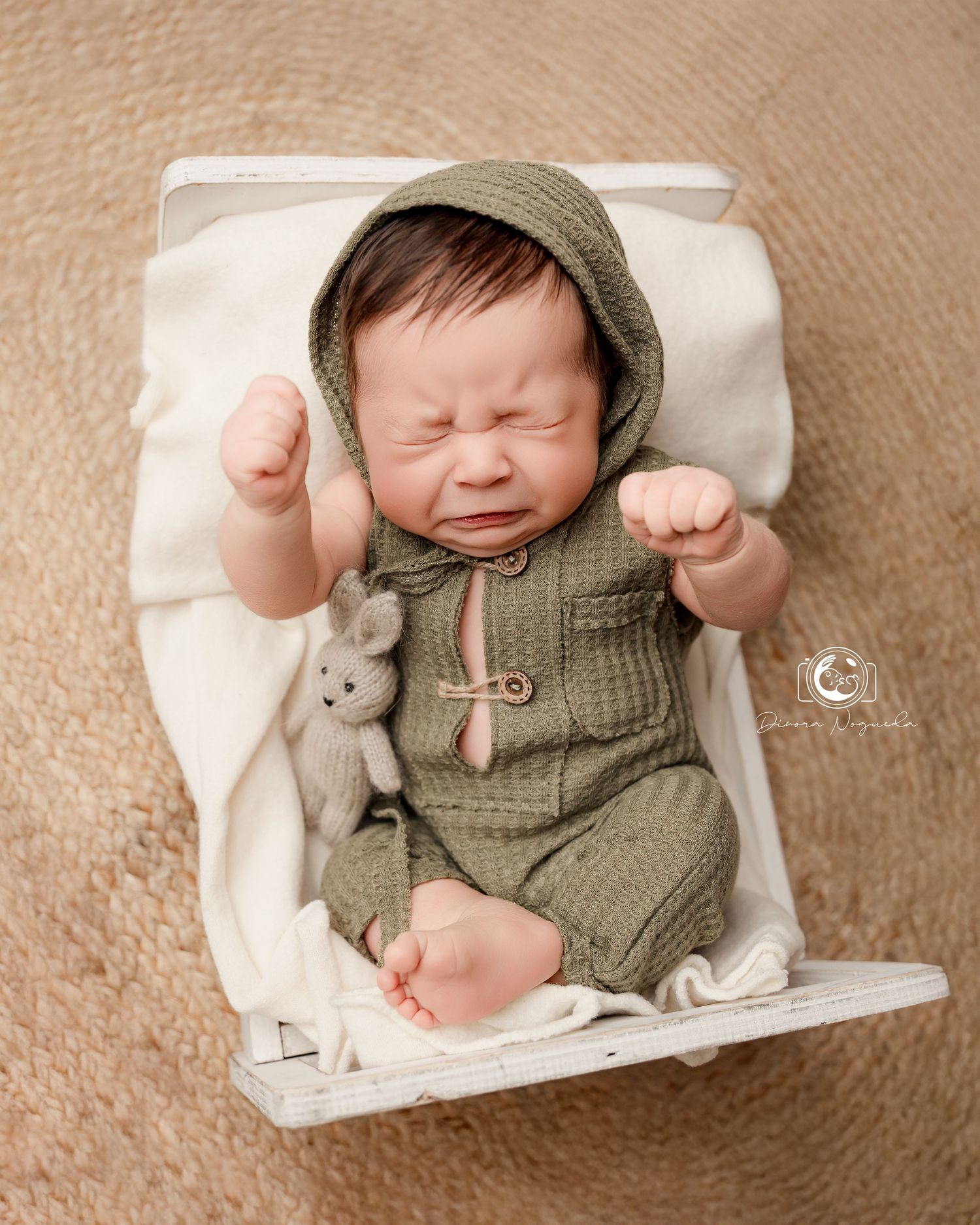 A baby wearing a knit olive green romper and beanie lies on a cream-colored blanket for a newborn photoshoot.