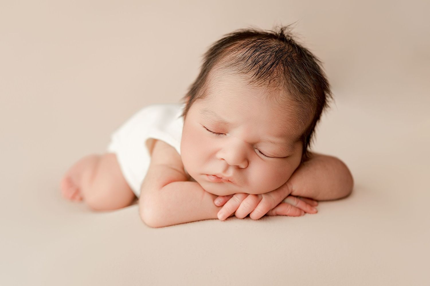 Peaceful newborn baby sleeping on cream backdrop wearing white onesie, tiny hands folded under cheek.