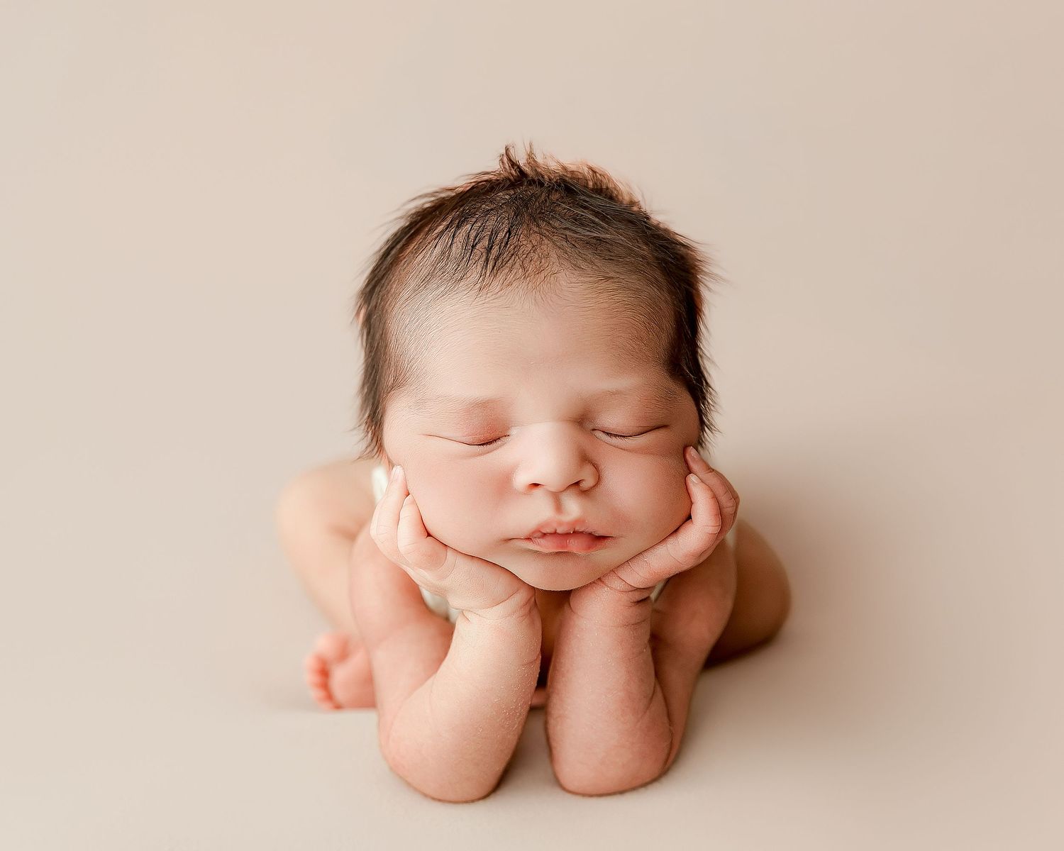 Peaceful newborn baby sleeping with hands under chin in classic froggy pose on beige backdrop.