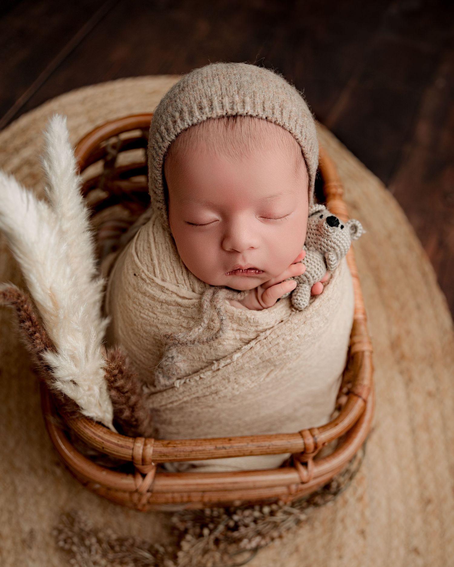 Sleeping newborn in knit bonnet and wrap posed in woven basket with white feather accent.