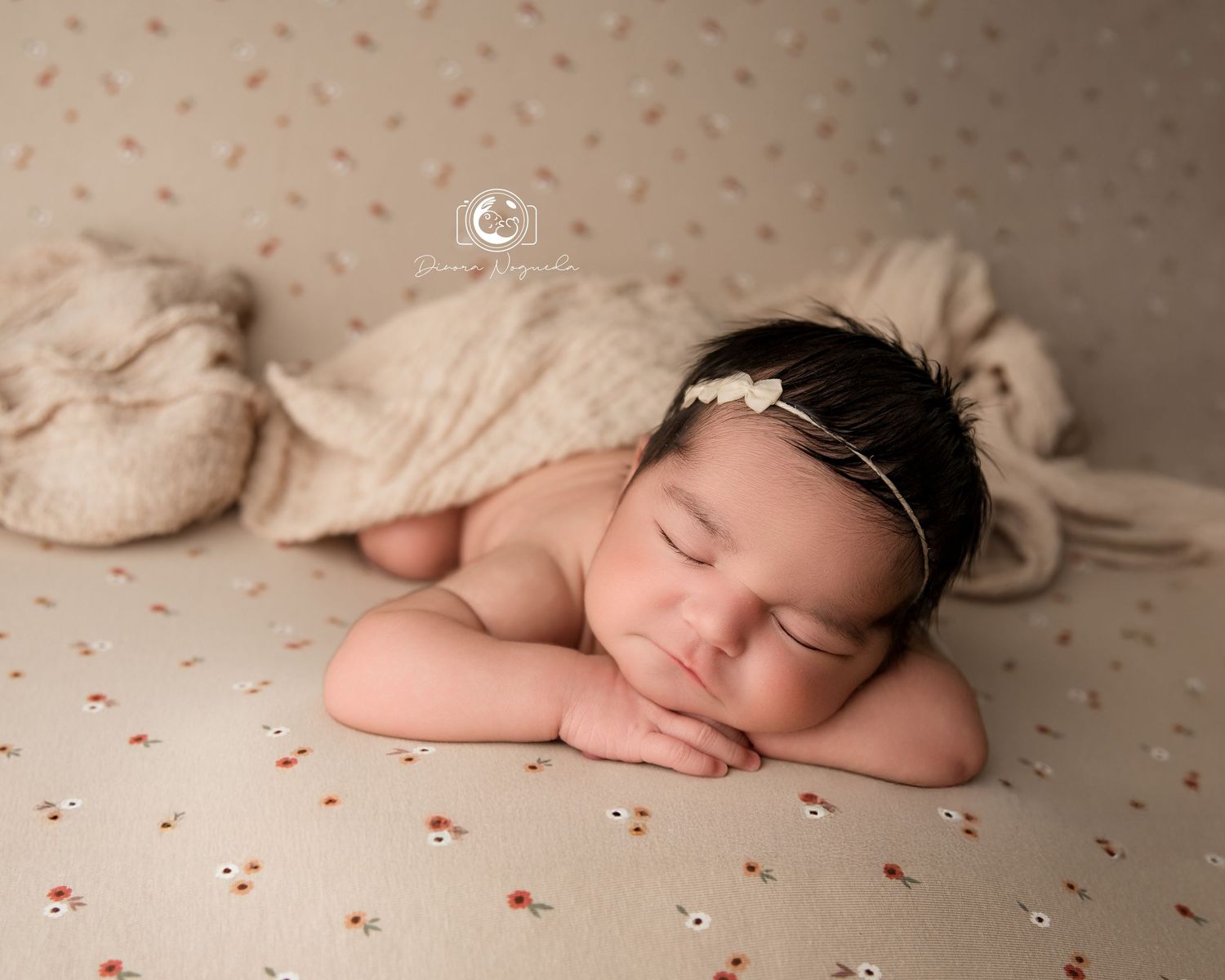 Peaceful sleeping newborn baby posed on soft beige fabric with delicate pink floral pattern and pearly headpiece.