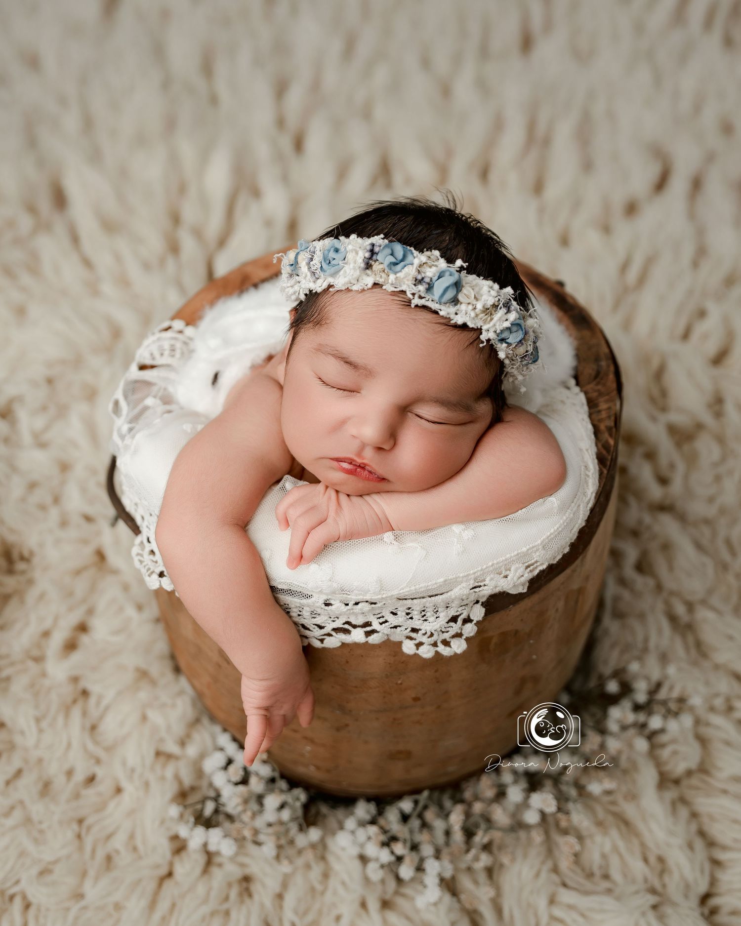 Sleeping newborn rests peacefully in a rustic wooden bowl adorned with delicate lace and floral headpiece.