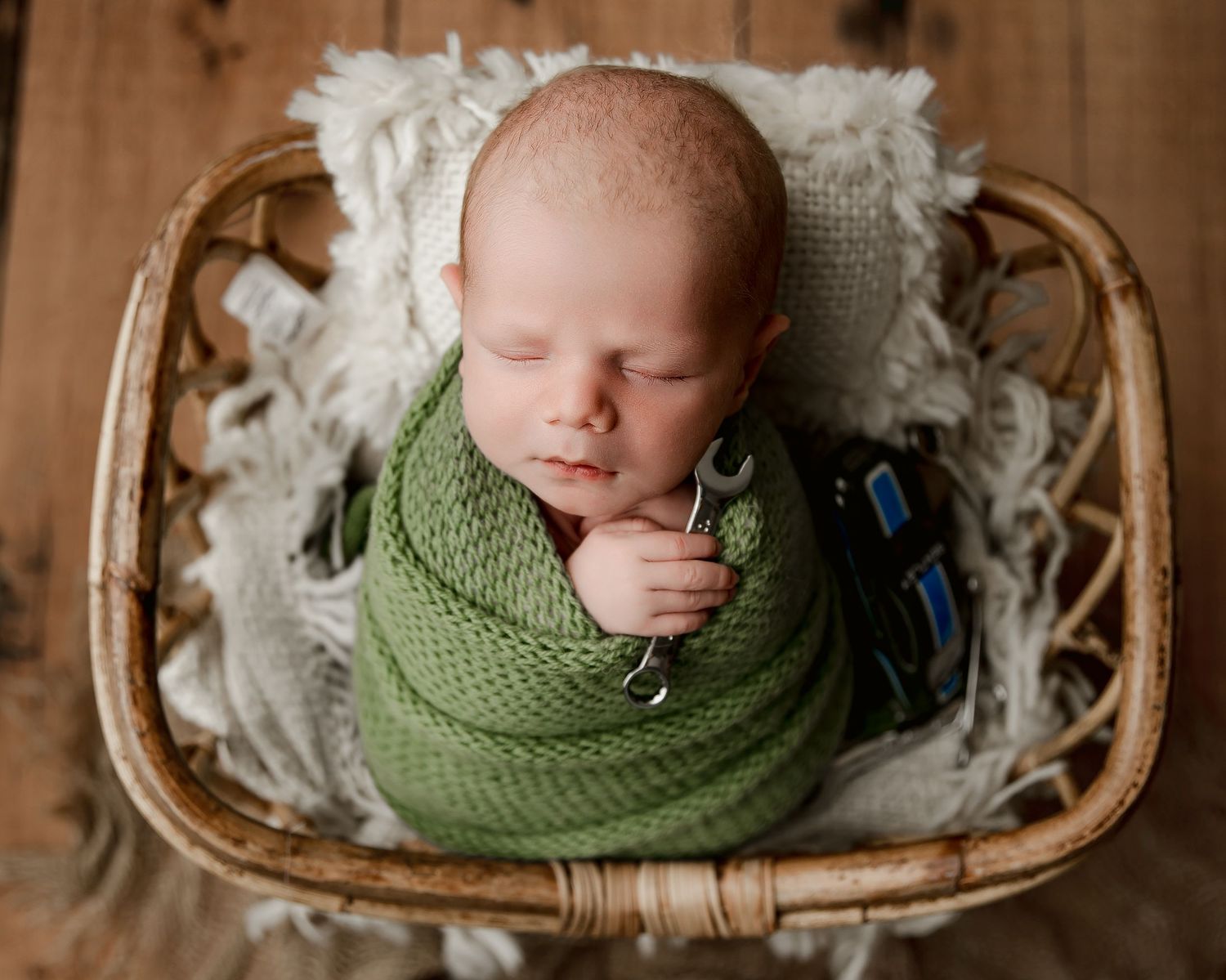 Newborn baby swaddled in green knit wrap holding a wrench, sleeping peacefully in a wicker basket with white fur.