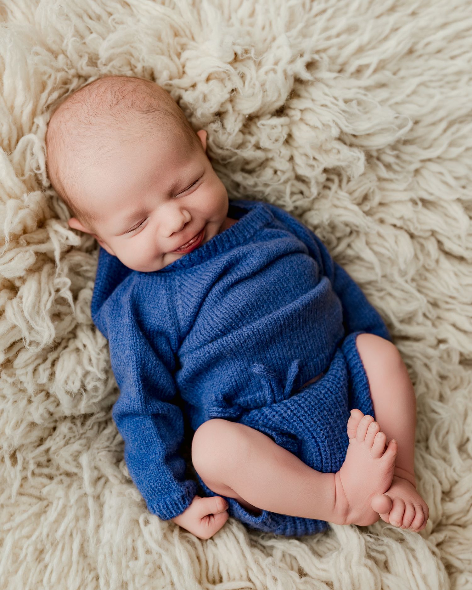 Smiling newborn baby in blue knit romper lying on fluffy cream rug.