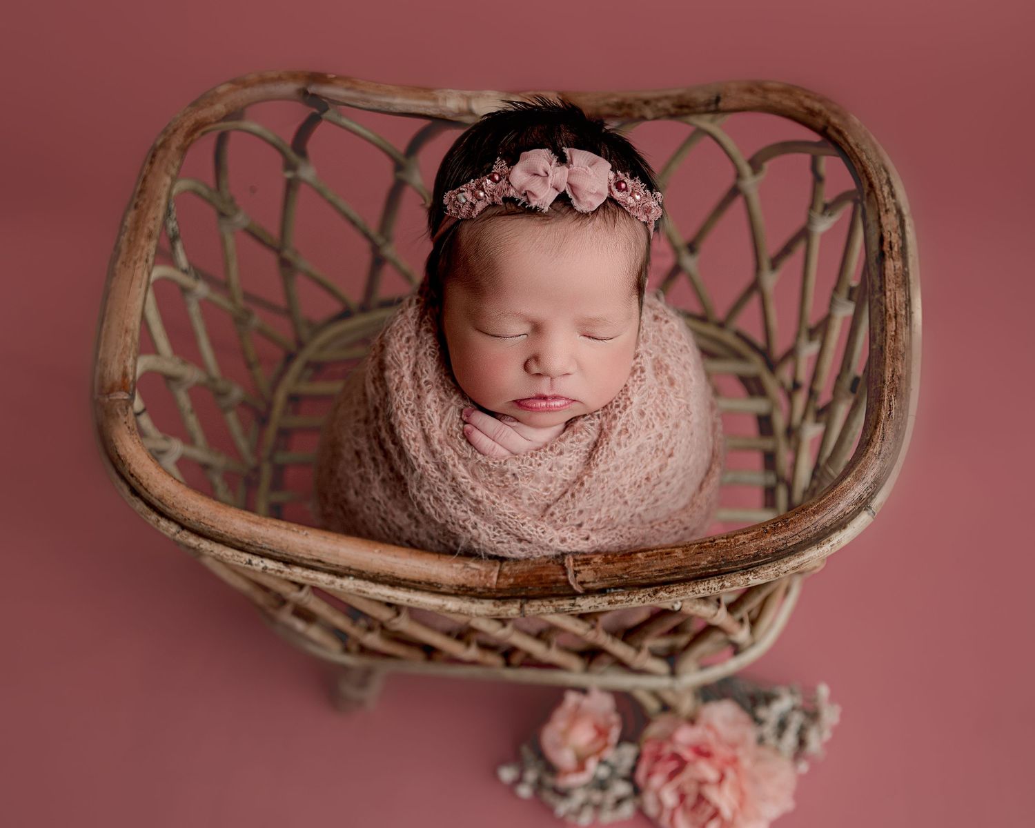 Peaceful newborn swaddled in pink wrap sleeping in vintage basket with floral accents.