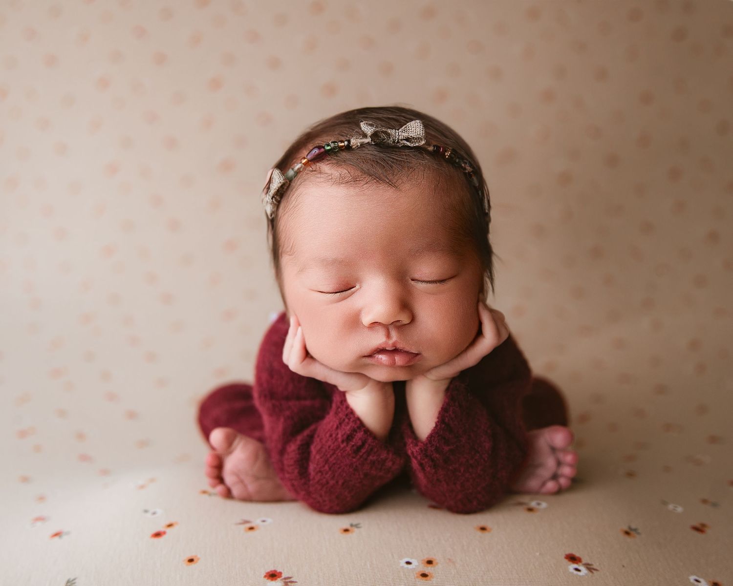 Serene sleeping baby in burgundy outfit with hands under chin on dotted background.
