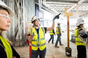 Construction workers in safety vests and hard hats discuss plans on a building site, with one pointing upward.