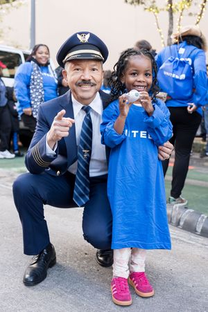 A pilot in uniform kneels beside a smiling young girl in a blue United shirt, pointing at the camera outdoors.