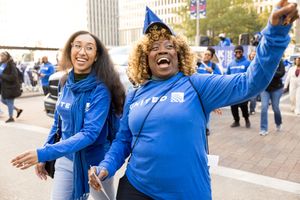 Two women in blue United hoodies celebrate at an outdoor rally, one cheering with arms wide open.