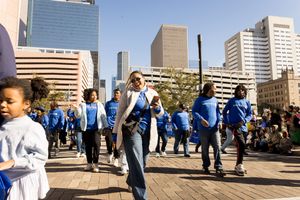 Group of youth in blue shirts performing at an outdoor urban parade with city skyscrapers in the background.