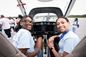 Two young women smile in the cockpit of a small aircraft at an aviation event outdoors.