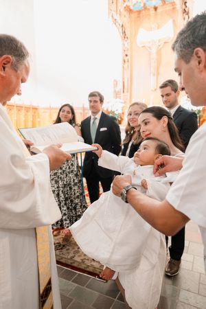 Catholic priest performing infant baptism ceremony with family gathered around altar in ornate church.