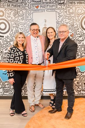Four people cutting an orange ribbon at a grand opening ceremony in front of a colorful mural.