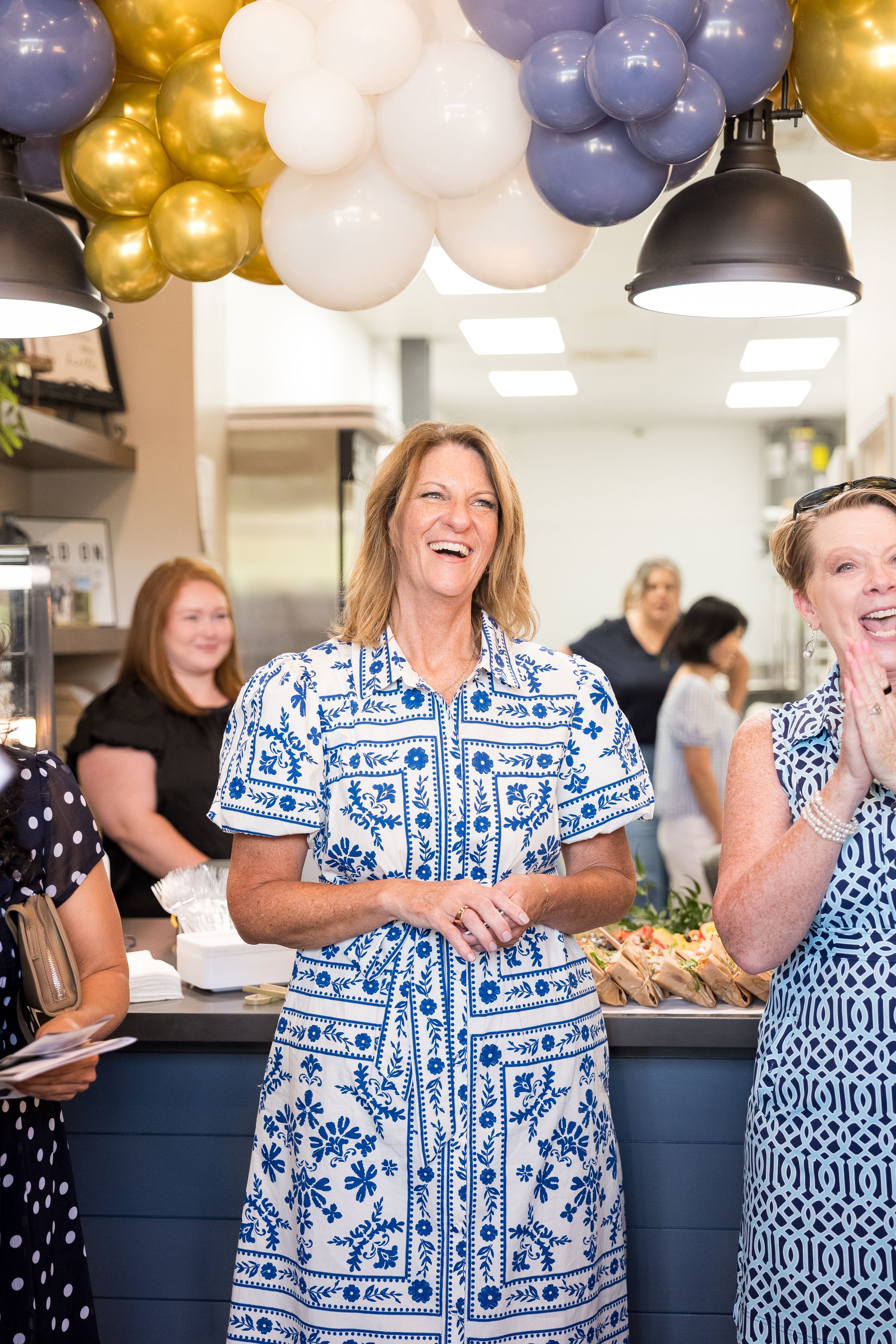 Woman in blue floral dress laughing at a celebration event with gold and purple balloons in a cafe.