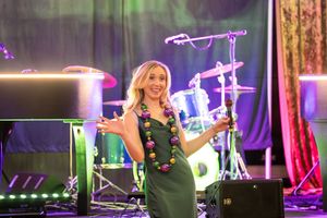 Woman in green dress and colorful lei smiling on stage with drums and keyboard in background.