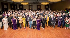 Large group of elegantly dressed people posing together at a formal gala event in a grand ballroom.