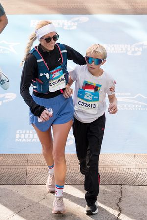 Two runners wearing race bibs compete in a marathon, with an adult guiding a young child toward the finish line.