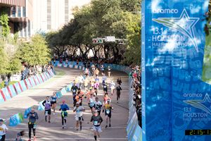 Runners compete in the Aramco Houston Half Marathon race through city streets with a digital clock display showing race time.