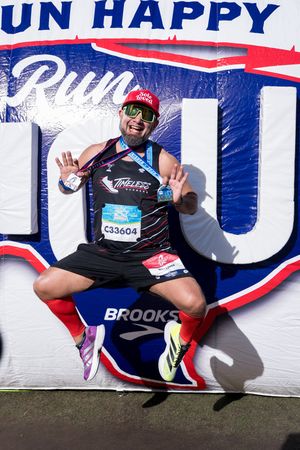 Excited marathon runner jumps in front of Run Happy banner wearing bib C33604 and finisher medal.