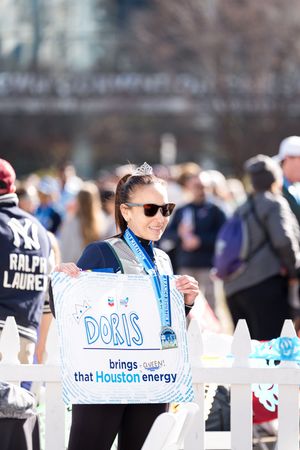 Woman named Doris holds a sign saying she brings Houston energy, wearing a tiara and medal at a marathon event.