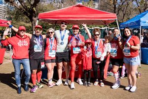 Group of marathon runners posing with medals under a red tent at an outdoor race event on a sunny day.