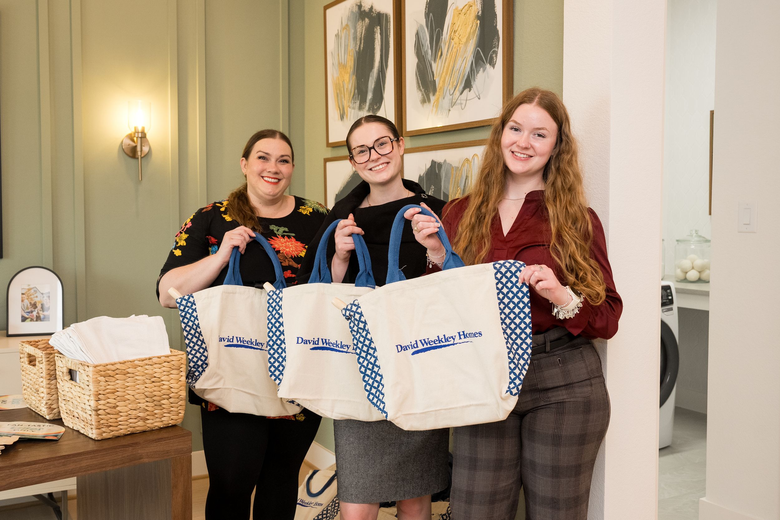 Three women smiling and holding David Weekley Homes branded tote bags inside a model home.