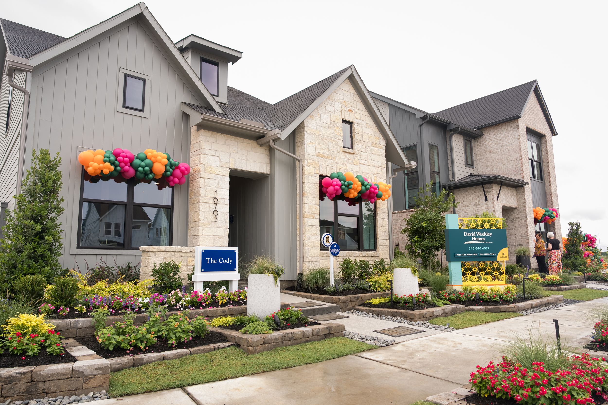 Colorful balloon-decorated model homes with stone and gray siding facades, manicured gardens, and vibrant flowers on a sunny day.