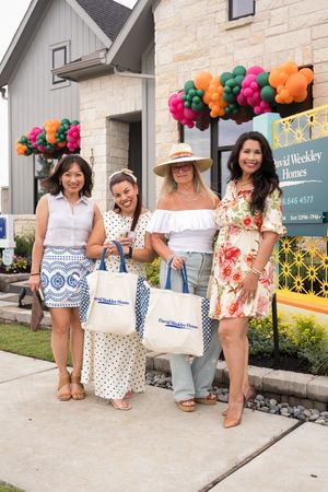 Four women pose with David Weekley Homes tote bags outside a model home decorated with colorful balloon garlands.