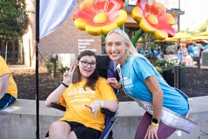 Two smiling women in yellow and blue charity event t-shirts pose together outdoors, one seated in a wheelchair.