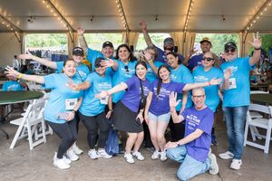 Group of volunteers in blue and purple t-shirts posing cheerfully under a tent at an outdoor charity event.