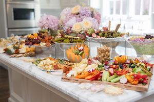 Elegant grazing table with charcuterie, fresh vegetables, fruits, and floral arrangements on a marble kitchen island.