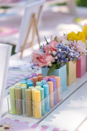 Colorful chalk pastels in clear organizer beside fresh pink and purple flowers on a white table.