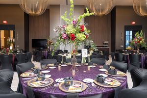 Elegant banquet table with purple sequin linen, floral centerpiece, and black chairs in a modern event venue.