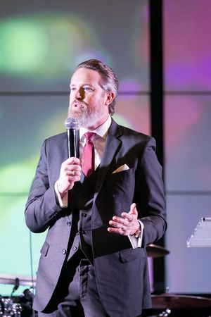 Man in gray suit with red tie speaks into microphone on stage with colorful lights behind him.