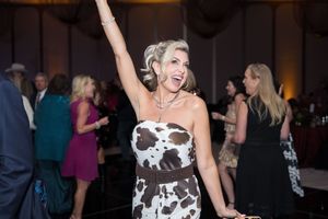 Woman in cow-print dress celebrating at elegant event, raising arm joyfully among guests in ballroom.