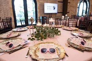 Elegant wedding reception table with gold chargers, pink sunglasses favors, greenery centerpiece, and candles in a brick venue.