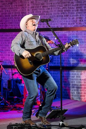 Country musician in white cowboy hat plays acoustic guitar and sings joyfully on stage with colorful brick backdrop.