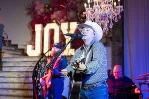 Country musician in white cowboy hat plays guitar on stage with JOY sign and chandelier backdrop.