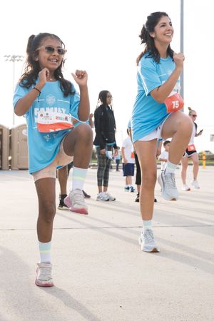 Two women in blue shirts do high knees warm-up exercises at a charity fun run event outdoors.