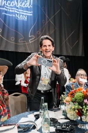 Man smiling and holding up a phone at a NAN Foundation Uncorked fundraising gala event.