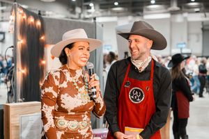 Two people in cowboy hats, one holding a microphone, smile at an indoor event venue.