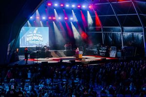 Live concert stage with colorful lights, large screen, and crowd at an outdoor reunion music festival event.