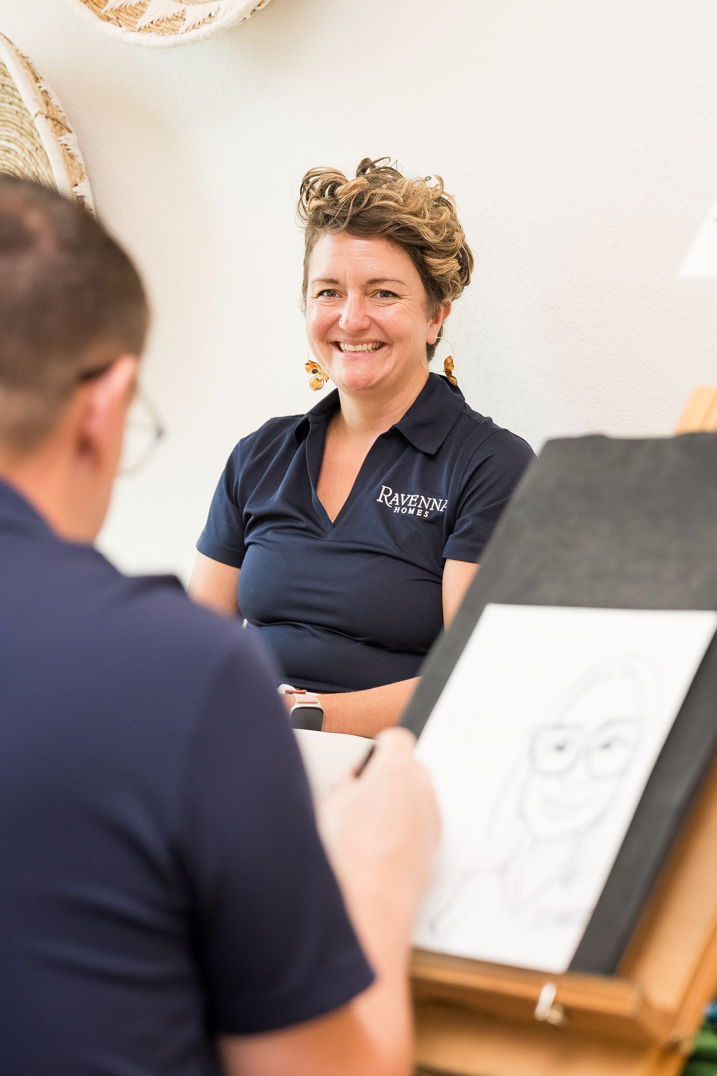Woman in Ravensbar polo shirt smiling while artist sketches her caricature portrait at an event.