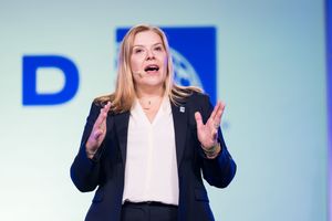 Professional woman speaker presenting on stage with blue corporate backdrop, wearing blazer and gesturing confidently.